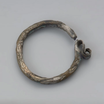 kabłączek skroniowy - Ujęcie dołu z góry. Brązowy kabłączek skroniowy z litego metalu z esowatym spłaszczonym uszkiem podwójnie zwiniętym w spiralę. Powierzchnia spatynowana, ciemnobrązowa.