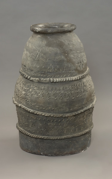 pojemnik; naczynie zasobowe - Ujęcie z przodu. Częściowo zrekonstruowane naczynie zasobowe o workowatym kształcie. Powierzchnia naczynia jest ornamentowana czterema strefami z motywami zdobniczymi oddzielonymi trzema dookolnymi listwami plastycznymi, na których znajdują się szerokie ukośne nacięcia przypominające karby. Pierwszą strefę przyozdabiają cztery rzędy niestaranie wykonanych, dookolnych linii falistych. Tuż pod falami znajdują się trzy rzędy lewo-skośnych nacięć. Drugą i trzecią strefę zdobią po cztery rzędy lewo-skośnych nacięć. Zasobowiec wylepiono z glinianych wałków z masy garncarskiej wzbogaconej domieszką piasku oraz granitu o różnorodnym stopniu granulacji. Naczynie wylepiono najprawdopodobniej ręcznie bez użycia koła garncarskiego.