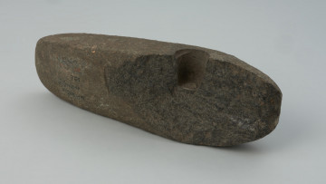 półwytwór, topór - Ujęcie z tyłu. Półwytwór topora kamiennego z prostym korpusem, pękniętym w trakcie wiercenia otworu w części tylnej. Powierzchnia po wstępnej obróbce ściernej. Otwór wiercowny z obu stron, pogłębiany od jednej. W przekroju otworu zaczątkowego widoczny fragment czopu powstającego przy wierceniu pustym wiertłem