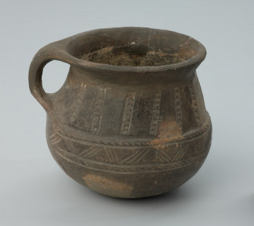 Gliniane naczynie (niewielki kubek z uchem) - Ujęcie prawego boku. Gliniane naczynie (niewielki kubek z uchem) o gładkiej powierzchni zewnętrznej barwy ciemnobrunatnej.