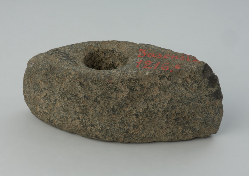 Topór kamienny - Ujęcie z lewego boku. Topór o wydłużonej części tylnej zakończonej wąskim obuchem, z wywierconych otworem przesuniętym ku tyłowi. Przekrój poprzeczny czworokątny wysoki. W w rzucie bocznym trapezowaty z lekko skośnym ostrzem i lekko łukowatym obuchem. Przy otworze, na stronie górnej, ślady wykucia płytkiej miseczki pod wiercenie. Powierzchnia zerodowana ze śladami szlifowania i puncowania. Ostrze wtórnie uszkodzone