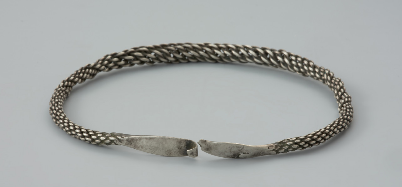 naszyjnik srebrny - Ujęcie zapięcia naszyjnika. Srebrny naszyjnik spleciony z pięciu par drutów owiniętych wokół siebie, z końcami rozklepanymi w owalne, niezdobione płytki. Jedna z nich zakończona została haczykiem, końcówka drugiej jest ułamana. W części środkowej druty są skręcone luźniej, co powoduje, ze obręcz jest w tym miejscu grubsza, natomiast po obu stronach znajdują się fragmenty, gdzie splot jest nierówny.