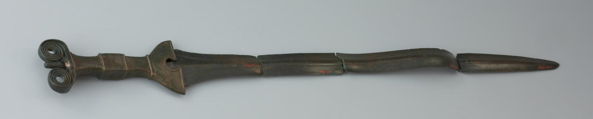 Miecz antenowy - Ujęcie miecza z tyłu. Miecz antenowy o wąskiej głowni, zachowanej w czterech częściach. Po obydwu stronach głowni biegną delikatne, podwójne strudziny. Czworokątny trzon rękojeści u podstawy ma dzwonowaty jelec. Głowica zaopatrzona jest w parę czterozwojowych ślimacznic. Krawędzie rękojeści zdobią drobne nacięcia.