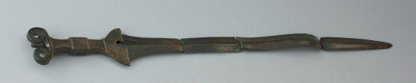 Miecz antenowy - Ujęcie miecza z tyłu. Miecz antenowy o wąskiej głowni, zachowanej w czterech częściach. Po obydwu stronach głowni biegną delikatne, podwójne strudziny. Czworokątny trzon rękojeści u podstawy ma dzwonowaty jelec. Głowica zaopatrzona jest w parę czterozwojowych ślimacznic. Krawędzie rękojeści zdobią drobne nacięcia.