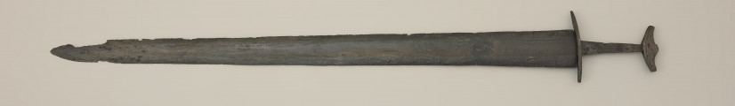 miecz dwusieczny - Ujęcie z tyłu. Całkowicie zachowany, z niewielkimi ubytkami korozyjnymi w okolicach sztycha. Jego głownia jest najszersza przy jelcu i równomiernie zwężą się ku ostremu sztychowi. Jest zaopatrzona w szerokie zbrocze. Głownię wykonano w technice prętowej, poprzez zgrzanie stalowych nakładek ostrzowych z jednolitym rdzeniem. Trzpień mocno zwęża się ku głowicy. Jelec, soczewkowaty w rzucie z góry, jest długi i płaski, a jego końce są zakrzywione w kierunku głowni. Jednolita głowica ma kształt siodełkowaty. Jej dolna część wygięta jest ku górze. Na jej powierzchni zaznaczono symboliczny podział na podstawę i nakładkę.