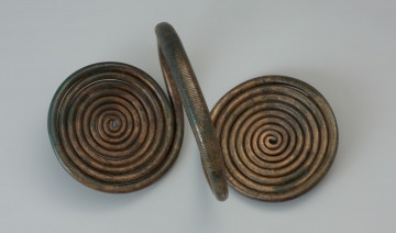 Naramiennik z tarczkami spiralnymi typu Czernice - Ujęcie spodu naramiennika. Naramiennik z brązowego drutu, wygięto w kabłąk, tworząc obręcz. Końce drutu uformowane zostały w dwie spiralne tarczki po obu stronach obręczy i jednocześnie prostopadle do niej. Grupy poprzecznych kresek oraz motywy jodełkowe tworzą ornament na kabłąku, a ukośne i poprzeczne nacięcia zdobią tarczki.