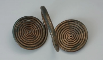 Naramiennik z tarczkami spiralnymi typu Czernice - Ujęcie spodu naramiennika. Naramiennik z brązowego drutu, wygięto w kabłąk, tworząc obręcz. Końce drutu uformowane zostały w dwie spiralne tarczki po obu stronach obręczy i jednocześnie prostopadle do niej. Grupy poprzecznych kresek oraz motywy jodełkowe tworzą ornament na kabłąku, a ukośne i poprzeczne nacięcia zdobią tarczki.