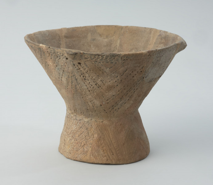Naczynie gliniane - Ujęcie lewego boku; Puchar na pustej nóżce, o klepsydrowatej formie, zdobiony na całej powierzchni ornamentem kreskowo-kłutym wykonanym dwuzębnym narzędziem: na czaszy – w układzie wiszących, wsuwanych kątów, które górą dochodzą do dookolnego pasma linii wykonanych tą samą techniką, a na nóżce – ukośnymi, zbieżnymi pasami linii. Przy krawędzi wylewu znajduje się zdobienie plastyczne w postaci czterech drobnych guzków