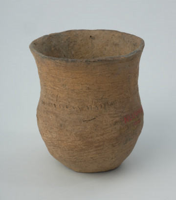 Naczynie gliniane - Ujęcie z przodu; Puchar o łagodnie esowatym kształcie, zdobiony na szyjce dookolnym pasmem odcisków sznura złożonym z 6 rzędów o nieumiarowym (falującym) przebiegu (ostatnie, najniższe pasmo jest przerwane w jednym miejscu) podkreślonym u dołu dookolną linią przecinkowatych odcisków, z brzuścem chropowaconym na powierzchni zewnętrznej. Dno uszkodzone (brak powierzchni zewnętrznej). Naczynie wyklejane (górna partia).