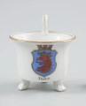 filiżanka w kształcie cylindrycznym - Ujęcie z przodu. Filiżanka z białej porcelany o kształcie cylindrycznym, rozszerzającym się ku górze, na trzech nóżkach w formie łap zwierzęcych. Uszko wolutowe wysunięte ponad czarkę w obrębie zawinięcia woluty przymocowanej dołem do krawędzi czarki. Kształt uszka podkreślony w środku i na bocznych krawędziach złotym paskiem. Na ściance podszkliwna dekoracja - barwny herb Szczecina - niebieska tarcza ze złotym obrzeżem, o ściętych narożnikach, zwieńczona sepiowo-brązową koroną muralis. Wewnątrz tarczy czerwona głowa gryfa z czarnym rysunkiem piór, ze złotą koroną. Krawędź czarki opracowana złotą lamówką, powtórzoną wąskim paskiem wokół wewnętrznej krawędzi czarki. Nóżki, na których opiera się czarka, wyprofilowane w poczwórne pazury podkreślone złotym akcentem.