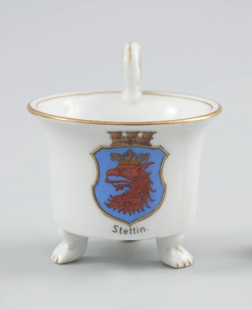 MNS/H/963/1/a filiżanka w kształcie cylindrycznym - Ujęcie z przodu. Filiżanka z białej porcelany o kształcie cylindrycznym, rozszerzającym się ku górze, na trzech nóżkach w formie łap zwierzęcych. Uszko wolutowe wysunięte ponad czarkę w obrębie zawinięcia woluty przymocowanej dołem do krawędzi czarki. Kształt uszka podkreślony w środku i na bocznych krawędziach złotym paskiem. Na ściance podszkliwna dekoracja - barwny herb Szczecina - niebieska tarcza ze złotym obrzeżem, o ściętych narożnikach, zwieńczona sepiowo-brązową koroną muralis. Wewnątrz tarczy czerwona głowa gryfa z czarnym rysunkiem piór, ze złotą koroną. Krawędź czarki opracowana złotą lamówką, powtórzoną wąskim paskiem wokół wewnętrznej krawędzi czarki. Nóżki, na których opiera się czarka, wyprofilowane w poczwórne pazury podkreślone złotym akcentem.