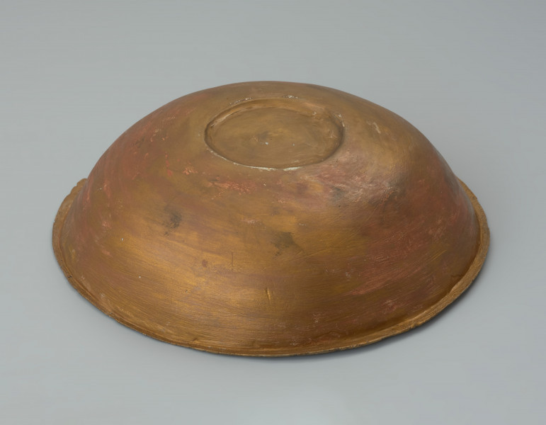 misa z rytem - Ujęcie spodu bokiem z odchyleniem do góry. Ozdobna misa wykonana została z jednego fragmentu blachy. Jej wąski, na zewnątrz wywinięty brzeg, miejscami opuszczony ku dołowi, ma zaokrągloną i lekko pogrubioną krawędź. Misa jest częściowo uszkodzona (jeden znaczny ubytek oraz kilkanaście mniejszych). Brakuje części ścianki od dna aż do wylewu. Pomimo ubytku i zniszczenia powierzchni zachowała się dekoracja ryta obejmująca wnętrze naczynia. Na nieco wklęsłym dnie widoczny jest duży medalion, z dodatkowo wydzieloną za pomocą podwójnej linii rytej częścią centralną wypełnioną liniami łamanymi. Na wewnętrznej powierzchni ścianki umieszczono symetrycznie cztery owalne medaliony z podobnym motywem, a pomiędzy nimi cztery 