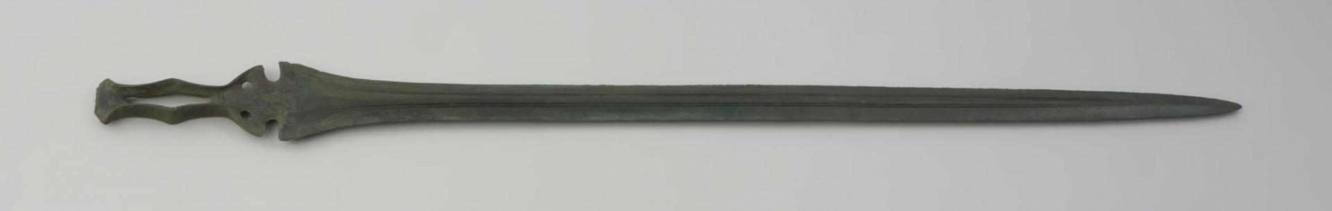 miecz ze sztabą do rękojeści - Ujęcie całego miecza w poziomie z tyłu. Zachowany w całości miecz obosieczny o smukłej głowni z widocznymi podwójnymi struzinami na powierzchni. Sztaba do rękojeści ma oryginalną formę, poszerzoną pośrodku i zwieńczoną wachlarzowatym zakończeniem. Dodatkowo na całej jej długości przebiega charakterystyczna, rombowata szczelina. Pókolista podstawa sztaby zaopatrzona jest w 2 otwory na nity. Obydwie powierzchnie miecza są lekko porowate i pokryte zieloną patyną.