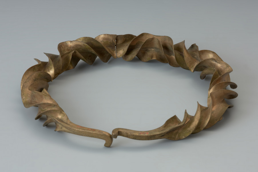 Naszyjnik wieńcowaty typu Wendelring - Ujęcie tyłu naszyjnika. Naszyjnik wieńcowaty charakteryzują szerokie, sfałdowane skrzydełka o ostrych krawędziach. Uzyskano je poprzez rozkuwanie i długotrwałe rozklepywanie pręta, pierwotnie o kwadratowym przekroju. Był skręcany wokół własnej osi przy kilkukrotnej zmianie kierunku skrętu. Czworokątne zakończenia naszyjnika ozdobione ornamentem punktowym wieńczy haczykowate zapięcie. Zabytek zachował się w dwóch częściach