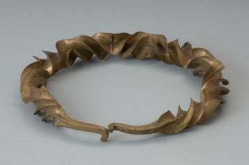 Naszyjnik wieńcowaty typu Wendelring - Ujęcie tyłu naszyjnika. Naszyjnik wieńcowaty charakteryzują szerokie, sfałdowane skrzydełka o ostrych krawędziach. Uzyskano je poprzez rozkuwanie i długotrwałe rozklepywanie pręta, pierwotnie o kwadratowym przekroju. Był skręcany wokół własnej osi przy kilkukrotnej zmianie kierunku skrętu. Czworokątne zakończenia naszyjnika ozdobione ornamentem punktowym wieńczy haczykowate zapięcie. Zabytek zachował się w dwóch częściach