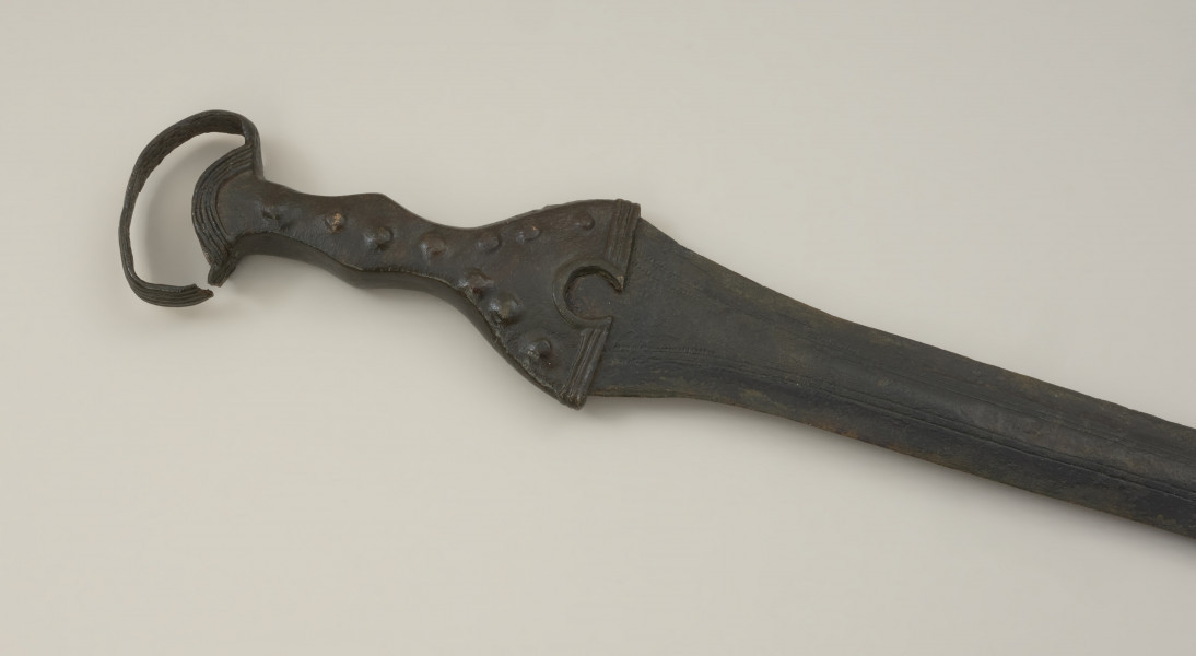broń - Ujęcie przodu rękojeści miecza w poziomie. Brązowy miecz posiada czworokątną rękojeść z nerkowatym pałąkiem oraz dzwonowaty jelec.