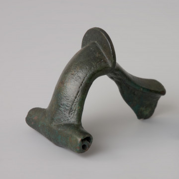 Zapinka z brązu - Ujęcie z przodu z lewej strony. Zapinka z brązu z kapturkiem pokrywającym od góry sprężynkę i plastycznym grzebykiem na kabłąku oraz zdobiona ornamentem rytym między grzebykiem a kapturkiem.