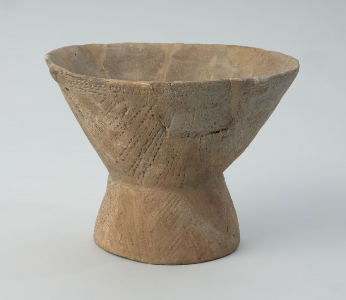 Naczynie gliniane - Ujęcie prawego boku; Puchar na pustej nóżce, o klepsydrowatej formie, zdobiony na całej powierzchni ornamentem kreskowo-kłutym wykonanym dwuzębnym narzędziem: na czaszy – w układzie wiszących, wsuwanych kątów, które górą dochodzą do dookolnego pasma linii wykonanych tą samą techniką, a na nóżce – ukośnymi, zbieżnymi pasami linii. Przy krawędzi wylewu znajduje się zdobienie plastyczne w postaci czterech drobnych guzków