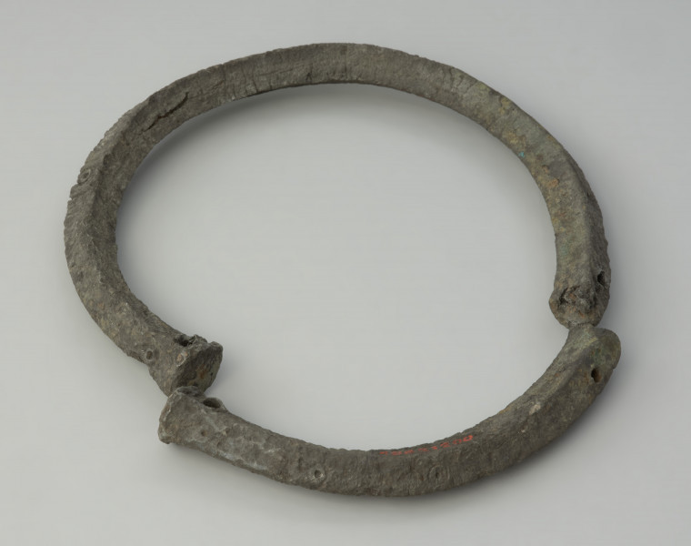 obręcz z czworograniastej sztaby - Ujęcie z góry tyłu obręczy. Srebrzystoszara obręcz, rozłamana na dwie części, wykonana została z czworograniastej cynowej sztaby. Przypomina kształtem naszyjnik obręczowy z ściętymi prosto i nieco pogrubionymi końcami oraz niewielkimi, nieregularnie rozmieszczonymi otworami. Zabytek jest zdeformowany, a jego powierzchnia popękana, co może świadczyć o wtórnym przepaleniu w ogniu.
