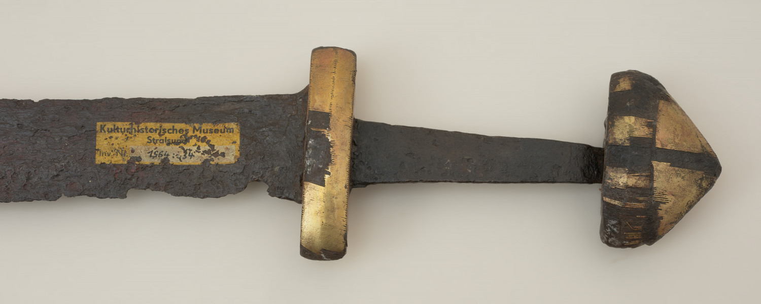 broń; miecz jednosieczny - Ujęcie rekojeści miecza z tyłu. Miecz o jednosiecznej głowni nieomal całkowicie zachowanej, z nielicznymi ubytkami korozyjnymi w partii sztychowej. Egzemplarz posiada nakładkę wysokowęglową nałożoną od strony ostrza na dwuwarstwową partię środkową. Trzpień mocno zwęża się ku głowicy. Jelec krótki i masywny z widoczną granią na bocznych powierzchniach, w rzucie z góry ma kształt łódkowaty. Głowica składa się z dwóch części. Jej podstawa podobna jest do jelca, a nakładka ma kształt trójkąta. Zarówno głowica jak jelec są zdobione w technice tauszowania (nabijania) z pasków stopu miedzi i cynku, ciasno spasowanych i wgrzanych w naciętą powierzchnię podkładu.