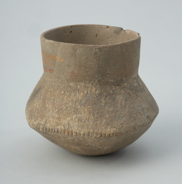 Naczynie gliniane - Ujęcie jednego boku;  Naczynie wazowate o dwustożkowatym brzuścu, pionowej szyjce i niewyodrębnionym dnie. Brzusiec o zarysie czworobocznym w rzucie z góry. Zdobione w górnej części brzuśca ukośnymi pasmami złożonymi z 3 podwójnych linii kreskowo-kłutych wykonanych dwuzębnym narzędziem. Pasma trzykrotnie zmieniają kierunek nachylenia, tworząc układ krokwiasty. Cały układ podkreślony dookolnym pasmem odcisków paznokcia na załomie brzuśca. Szyjka naczynia, niemal w całości zrekonstruowana, prawdopodobnie nie była zdobiona, podobnie jak dół brzuśca