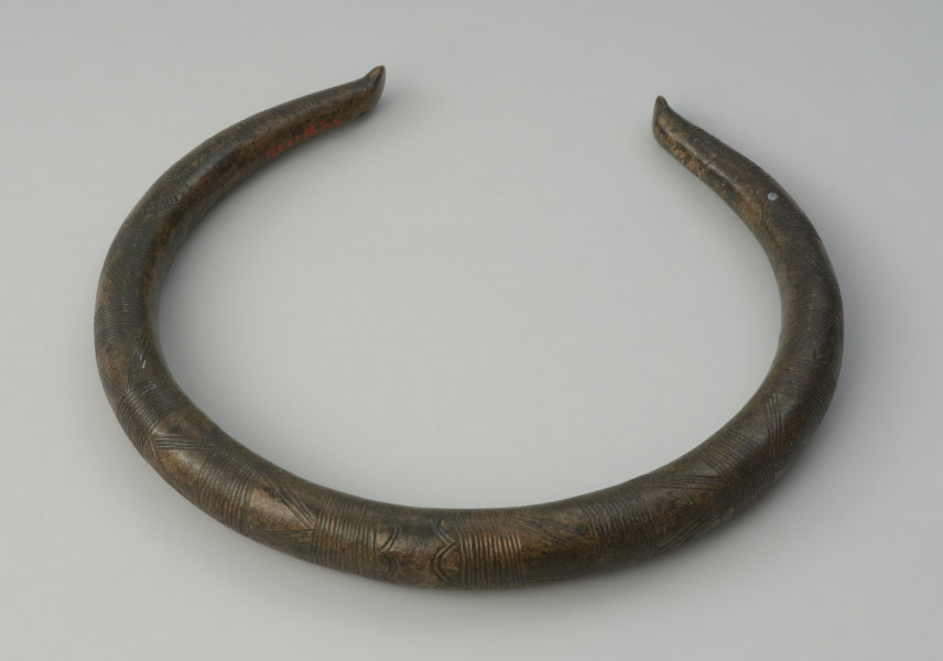 Brązowy naszyjnik - Ujęcie z góry. przodu naszyjnika. Brązowy naszyjnik wykonany z grubego okrągłego pręta. Jego zakończenia są odgięte na zewnątrz. Zdobiony jest poprzecznymi kreskami, trójkątami oraz łukami.