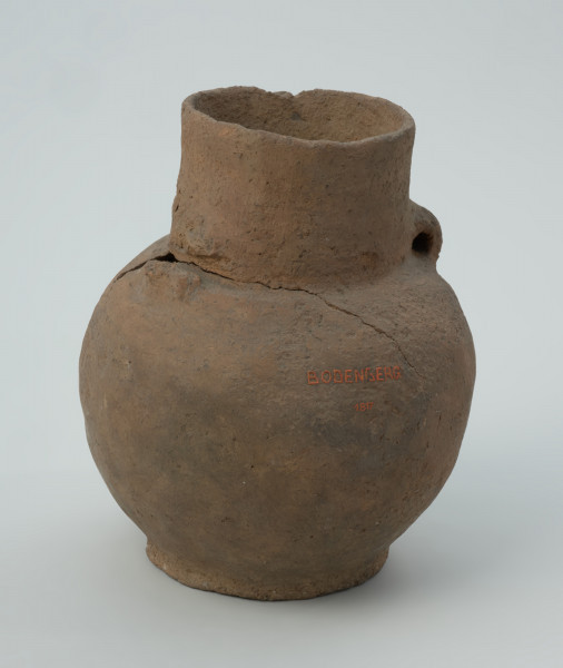 ceramika, naczynie - Ujęcie z przodu; Niezdobiona amfora z niemal cylindryczną szyjką, z parą taśmowatych, wątorowanych uszek na załomie szyjki i brzuśca. Z kulistym brzuścem i niską stopką. Uszkodzona przy przejściu szyjki w brzusiec (pęknięcie i brak ucha).