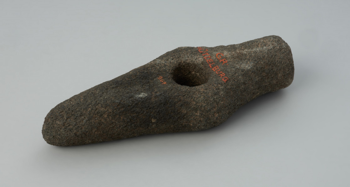 Topór kamienny - Ujęcie spodu; Topór kamienny z wklęsłymi powierzchniami górną i dolną oraz lekko wypukłymi powierzchniami bocznymi. Ostrze proste. Obuch tępym o zarysie kolistym. Korpus prosty w rzucie bocznym, w rzucie z góry z poszerzeniem przy otworze. Otwór kolisty, cylindryczny. Powierzchnia zerodowana, pierwotnie gładzona.