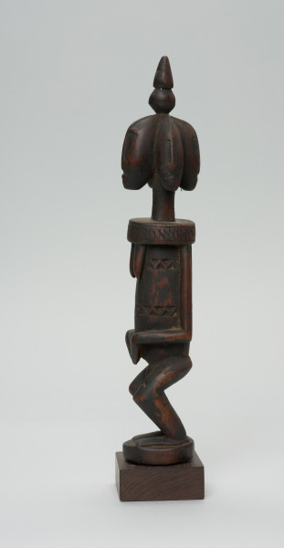 rzeźba - Ujęcie lewego boku. Drewniana, rzeźbiona postać z wyraźnie zaznaczonymi piersiami, a także z czterema głowami. Każda głowa zwrócona jest w inna stronę. Na czubku głowy stożkowata bryła. Twarze zaznaczone bardzo symbolicznie, nos w kształcie strzałki. Ramiona od szyi oddzielone prostą linią. Ręce przylegające do tułowia, dłonie złożone na łonie/podbrzuszu. Nogi krótkie, podkurczone w kolanach. Na brzuchu, na wysokości pępka dekoracyjne trójkątne nacięcia.