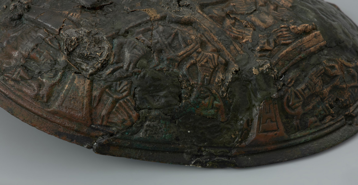 zapinka miseczkowata - Ujęcie fragmentu przedniej prawej części zapinki. Zapinka ma kształt wydłużonego owalu. Przedmiot jest częściowo uszkodzony bez zachowanej szpili. Powierzchnia fibuli jest bogato ornamentowana motywami zoomorficznymi. Na detal zdobniczy składa się osiem pól wyznaczonych przez krzyżujące się wstęgi. W punktach ich przecięcia znajdują się ślady po siedmiu wypukłych guzkach. Na sześciu z ośmiu pól znajdują się motywy zwierzęce bliskie skandynawskiej sztuce w stylu Borre. Na trzech z czterech zachowanych pól zlokalizowanych w narożnikach fibuli występują zwierzęta chwytające swoimi szponami ciała innych stworzeń. Dwa kolejne naprzeciwległe pola, usytuowane w środkowej części odpowiadają dwóm łukowato wygiętym w biodrach zwierzętom - być może są to konie z głowami skierowanymi w swoją stronę. Pozostałe dwa pola zawierają przedstawienia zwierzęcych masek z połączonymi ze sobą zaciśniętymi łapami.