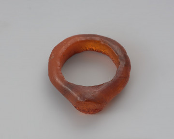 Jednoczęściowy pierścionek bursztynowy - Ujęcie z góry. Pierścionek bursztynowy z tarczką, jednoczęściowy, pomarańczowy, częściowo przejrzysty, częściowo mleczny. Jego otok jest w przekroju płaskokulisty i ma grubości ok. 0,4 cm. Powierzchnia zewnętrzna została wygładzona i nosi na sobie ślady obróbki, wewnętrzną natomiast pozostawiono nieobrobiona. Całość pokryta jest siatką drobnych pęknięć.