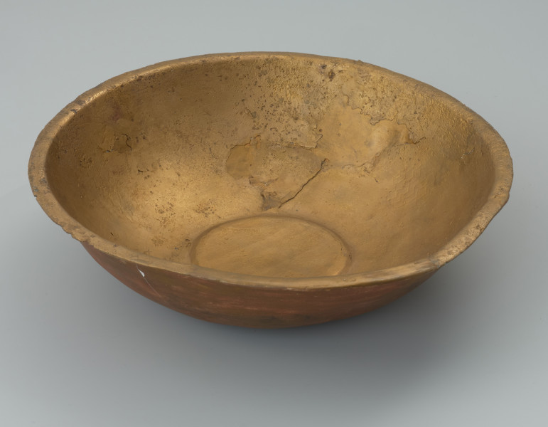 misa z rytem - Ujęcie z boku z odchyleniem do góry. Ozdobna misa wykonana została z jednego fragmentu blachy. Jej wąski, na zewnątrz wywinięty brzeg, miejscami opuszczony ku dołowi, ma zaokrągloną i lekko pogrubioną krawędź. Misa jest częściowo uszkodzona (jeden znaczny ubytek oraz kilkanaście mniejszych). Brakuje części ścianki od dna aż do wylewu. Pomimo ubytku i zniszczenia powierzchni zachowała się dekoracja ryta obejmująca wnętrze naczynia. Na nieco wklęsłym dnie widoczny jest duży medalion, z dodatkowo wydzieloną za pomocą podwójnej linii rytej częścią centralną wypełnioną liniami łamanymi. Na wewnętrznej powierzchni ścianki umieszczono symetrycznie cztery owalne medaliony z podobnym motywem, a pomiędzy nimi cztery 