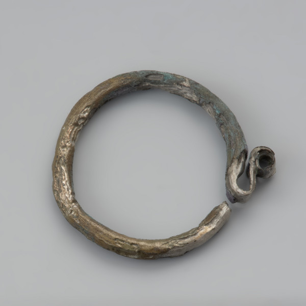 kabłączek skroniowy - Ujęcie przodu z góry. Brązowy kabłączek skroniowy z litego metalu z esowatym spłaszczonym uszkiem podwójnie zwiniętym w spiralę. Powierzchnia spatynowana, ciemnobrązowa.