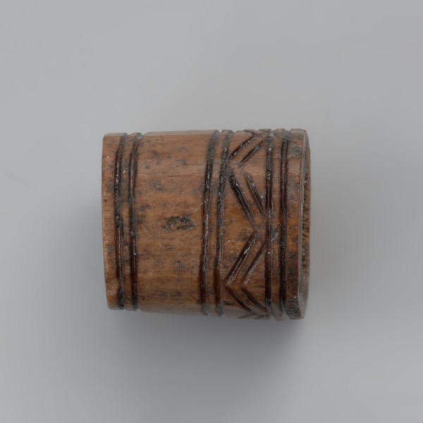 oprawka stożkowata - Ujęcie lewej strony w poziomie. Oprawka ma stożkowaty kształt o ściętym wierzchołku. Ślady pozostawione na jej powierzchni świadczą o wykorzystaniu do jej produkcji co najmniej trzech narzędzi – piły, noża oraz świdra. Na powierzchni oprawki znajduje się mało skomplikowany wzór zdobniczy o motywie liniowym naniesionym za pomocą ostrego narzędzia. Górną część zajmuje ornament w postaci dwóch dookolnych linii rytych. Natomiast na przeciwległej stronie występuje ozdobnik w formie linii zygzakowatych wpisanych od góry i od dołu w podwójne linie ryte.