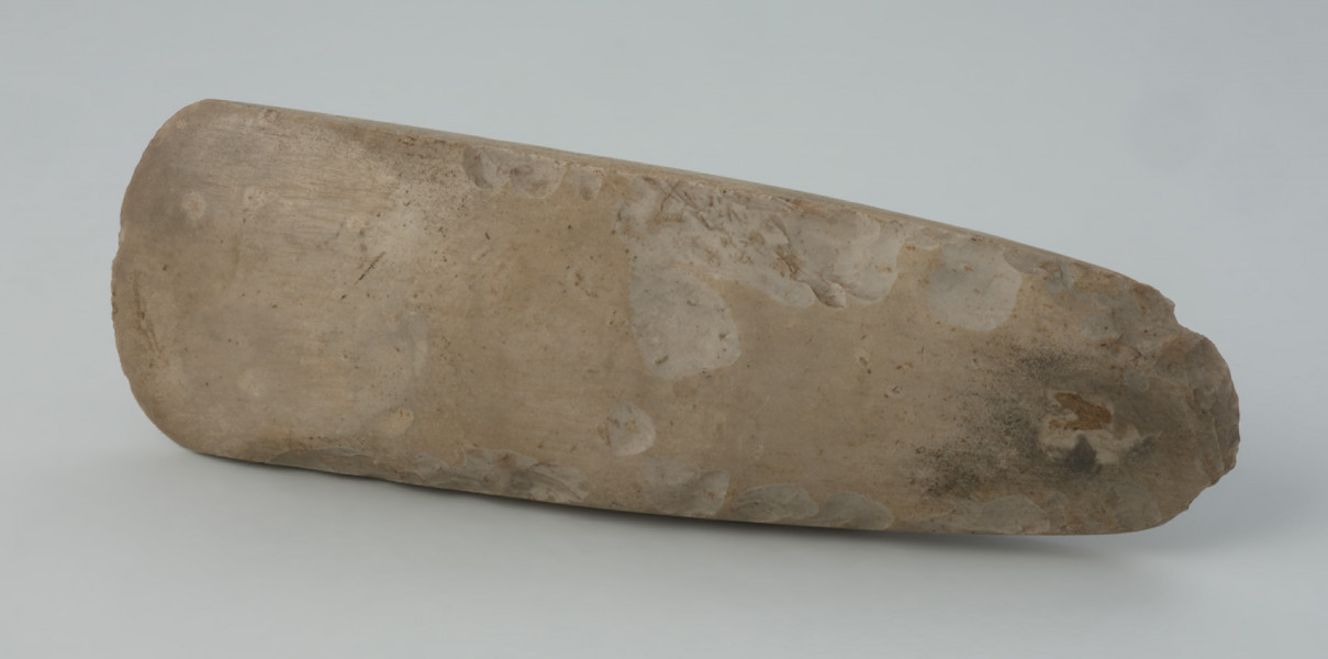 Siekiera krzemienna - Ujęcie z przodu; Długa, smukła siekierka czworościenna z cienkim obuchem, wykonana z krzemienia kredowego o niejednolitej, szarej barwie. Obuch i ostrze łukowe. Powierzchnia wygładzona, miejscami do połysku, ze śladami po rdzeniowaniu przy krawędziach bocznych i na obuchu. Na powierzchni szerszej ślad kory. Ostrze z drobnymi śladami wtórych uszkodzeń.