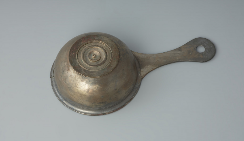 naczynie; Rzymski brązowy rondelek - Ujęcie z dołu. Rzymski brązowy rondelek z płaskim dnem o przewężonym uchwycie z otworem. Rondel jest zdobiony pod wylewem płytkimi dookolnymi liniami rytymi.