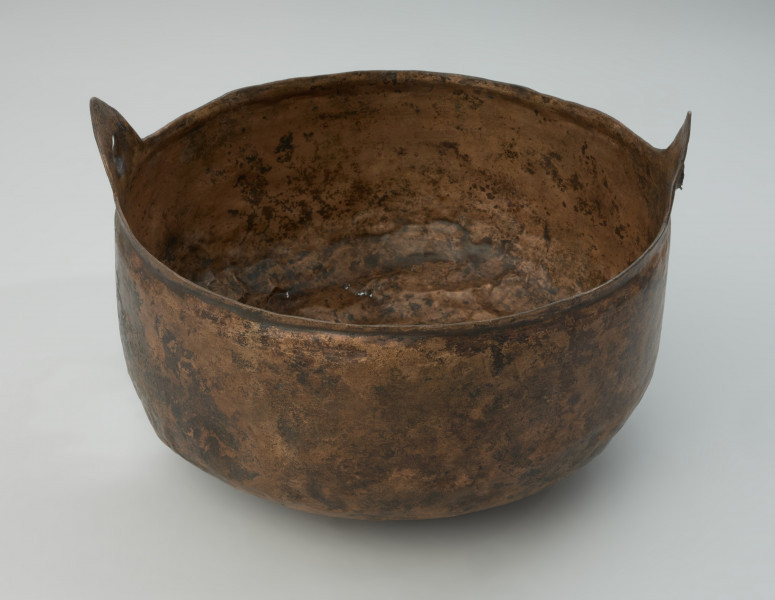 Kocioł ze stopu miedzi - Ujęcie z przodu. Kocioł ze stopu miedzi o owalnym kształcie z dwoma trójkątnymi uchami. Kocioł wykonano ze stopu miedzi. Jego kształt jest asymetryczny. W części górnej jest węższy i owalny, natomiast w części dolnej szerszy o zaokrąglonym dnie.