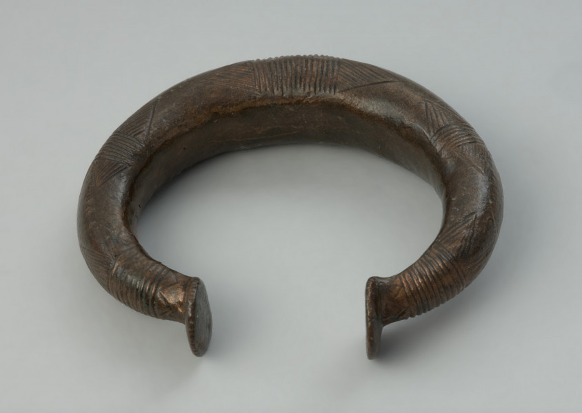 Bransoleta obręczowa - Ujęcie z góry tyłu. bransolety. Masywna bransoleta obręczowa o owalnym kształcie, odlana z brązu w formie grubego pręta. Jej zakończenia mają kształt niewielkich stopek. Powierzchnię obręczy zdobią grupy poprzecznych kresek oraz motyw tzw. wilczych zębów.