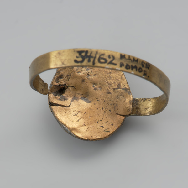pierścionek z oczkiem - Ujęcie tyłu pierścionka w poziomie. Trzyczęściowy brązowy pierścionek z oczkiem, składający się z metalowej taśmy stanowiącej otok, metalowej tarczki pod oczko złożonej z owalnej płaskiej blaszki spodniej oraz dodatkowej taśmy tworzącej eliptyczne gniazdko. Jest ona zdobiona dookoła odciskami. Trzecim elementem, który uzupełnia oczko jest grafitowe szkiełko zachowane jedynie w części, przyklejone do spodniej tarczki i przytrzymywane przez krawędzie otaczającej je taśmy. Między blaszkami oczka pozostawione są dwie 