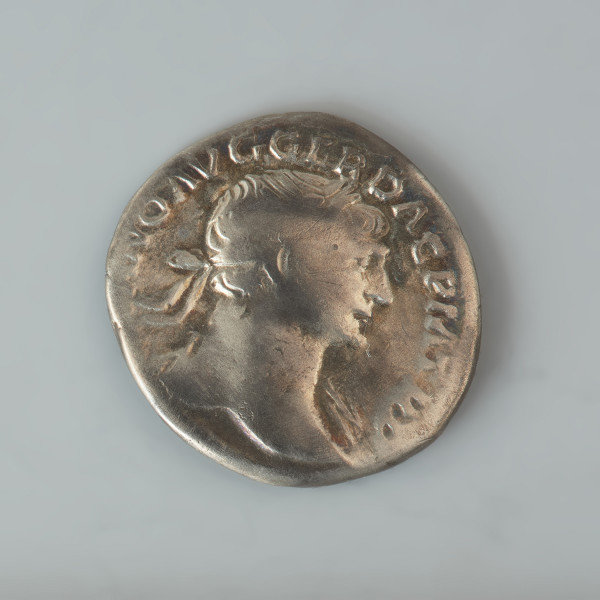 Ujęcie awersu. Denar Trajana. Powierzchnie są w znacznym stopniu starte, a wyobrażenia i napisy są częściowo nieczytelne. Na awersie widnieje popiersie cesarza w wieńcu laurowym, zwrócone prawym profilem, na lewym dalszym barku widoczna udrapowana szata. W otoku znajduje się napis IMP TRAIANO AVG GER DAC PM TRP. Na rewersie znajduje się personifikacja Dacii w żałobie lub wyobrażenie pokonanego Daka. Jest zwrócona w prawo postać w stroju dackim: spodniach, krótkiej tunice (płaszczu?) i szpiczastej czapce. Postać ta siedzi na stosie uzbrojenia prawa dłonią zakrywa twarz. Wokół wizerunku znajduje się częściowo tylko zachowany napis COS V PP SPQR OPTIMO PRINC.