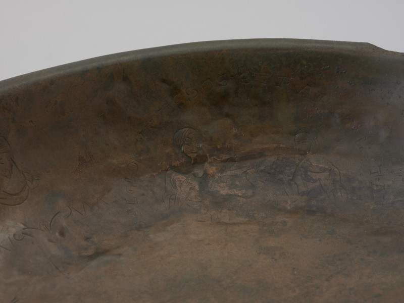 misa z rytem i tekstem - Ujęcie fragmentu wnętrza misy z jednym medialionem. Misa wykonana została ze stopu miedzi, z wąskim, wywiniętym, lekko pogrubionym brzegiem i płaskim dnem. Wewnętrzną powierzchnię ścianki zdobią cztery medaliony ze scenami z życia Heraklesa, otoczone łacińskimi inskrypcjami będącymi cytatami z eklogi przypisywanej Theodolusowi, które zapisano majuskułą romańską. Medaliony przedzielono wizerunkami koronowanych głów męskich.