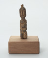 rzeźba - Ujęcie z przodu; Mała figurka antropomorficzna, najprawdopodobniej przedstawienie przodka. Głowa, rysy twarzy, ręce, nogi wydobyte z bryły drewna płytkimi, ostrymi, prostymi cięciami, co nadaje figurce zgeometryzowana formę. Mogła pełnić rolę osobistego fetysza.