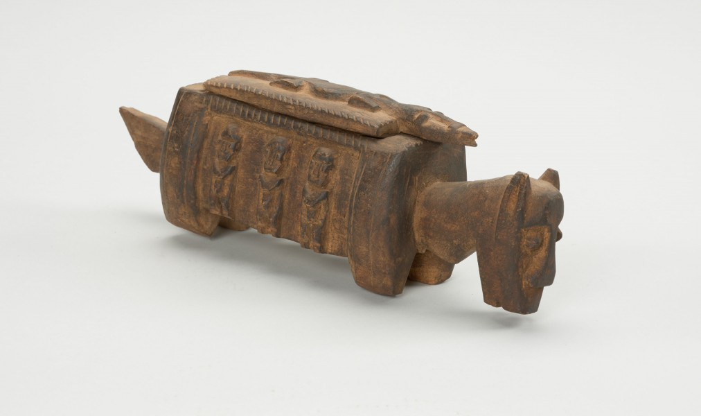 rzeźba - Ujęcie ze skosu z lewej; Arka Nommo zwana inaczej aduno koro lub arką świata, jest wyobrażeniem mitycznej łodzi. Drewniana rzeźa przypominająca konia. Bok zdobiony trzema postaciami w formie płaskorzeźby, stojące w rzędzie. Głowa obiektu zwrócona w stronę ziemi. Na grzbiecie druga część obiektu w formie przykrywki kształtem przypominająca krokodyla na drwnianej prostokatnej podstawie.