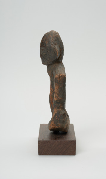 rzeźba - Ujęcie prawej strony. Figurka przedstawiająca postać ludzką. Ręce i nogi w przysiadzie wąskie, połączone ze sobą w formę kwadratu. Głowa spiczasta z odstającymi szerokimi uszami. Tułów cienki, prosty.