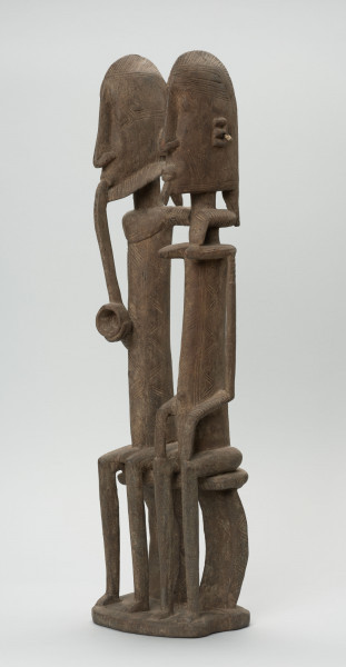 rzeźba - Ujęcie ze skosu z prawej; Figura - para przodków -  mężczyzny i kobiety. Oboje ujęci w pozycji siedzącej. Postacie smukłe. Mężczyzna w ustach ma fajkę, obejmuje lewą ręką kobietę. Związana z kultem przodków.