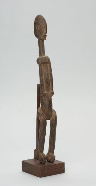 rzeźba - Ujęcie ze skosu z lewej; Figura - postać przodka. Figura antropomorficzna, sylwetka smukła, wydłużona. Ręce krótkie, dłonie ułożone nad pępkiem. Twarz schematyczna w stylu dogońskim. Szerokie biodra. Związana z kultem przodków.