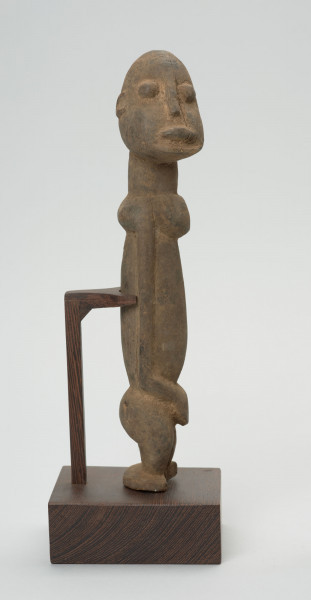 rzeźba - Ujęcie z lewego boku; Figura - postać przodka. Głowa i korpus w stosunku do nóg bardzo duże - co typowe jest dla sztuki dogońskiej. Na uwagę zasługuje przedstawienie twarzy, wykonane nie centralnie, ale z prawej strony. Co nadaje postaci cechy komiczne. I wprowadza do zwykle statycznych wizerunków trochę ruchu. Figura wspierana konstrukcją przymocowaną do podstawy.