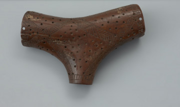 pojemnik trójdzielny - Ujęcie przodu pojemnika z góry. Pojemnik trójdzielny wykonany z wydrążonego fragmentu poroża jelenia posiada formę zbliżoną do litery T, wynikającą z naturalnego ukształtowania surowca. Każde z trzech okrągłych w przekroju ramion zaopatrzone jest w otwór. Na ich brzegach znajdują się po cztery dziurki służące mocowaniu zamknięć i zawiesi. Powierzchnia przedmiotu jest bogato zdobiona rytymi wstęgami oraz nieregularnie rozmieszczonymi wgłębieniami. Wstęgi wypełniono trójkątami ze szrafowaniem. Na stronie wklęsłej ornament wzbogacono krzyżem umieszczonym w nieregularnej obwódce. W jego centrum, u zbiegu ramion znajdują się cztery punkty-wgłębienia. Na odwrotnej stronie pojemnika umieszczono znak X.