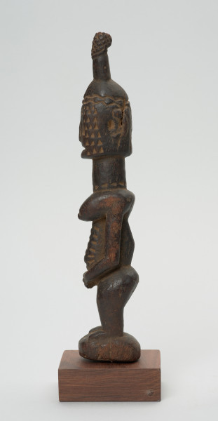 rzeźba - Ujęcie prawej strony rzeźby. Drewniana figura postaci kobiety. Podłużna głowa ze scheycznie zaznaczonymi rysami i ozdobną wysoką fryzurą. Długa masywna szyja, szczupły tułów. Wyraźnie zaznaczone piersi, długi ręce splecione z przodu. Postać ma krótkie nogi ugięte w kolanach.