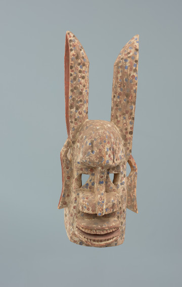 drewniana, rzeźbiona maska - Ujęcie z przodu. Drewniana, rzeźbiona maska, której część twarzowa jest podłużna, zaokrąglona na górze, oczy wycięte, trójkątne, czoło wypukłe, nozdrza i usta szerokie. Rogi proste, na końcach ścięte, uszy, opadające na dół. Maska pomalowana jest na biało, pokryta różowymi, niebieskimi, brązowymi kropkami.